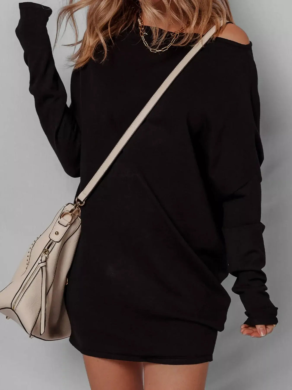 Woman wearing asymmetric batwing sweater dress with off-shoulder style, black color, paired with beige handbag.