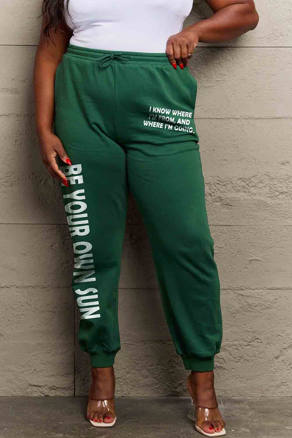 Woman wearing Graphic Sweatpants with 'BE YOUR OWN SUN' print, green color, drawstring waistband, full-size design.