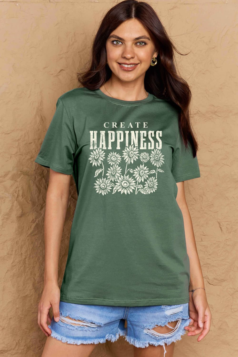 Woman wearing a green Graphic Cotton T-Shirt with 'CREATE HAPPINESS' slogan in floral print, paired with denim shorts.