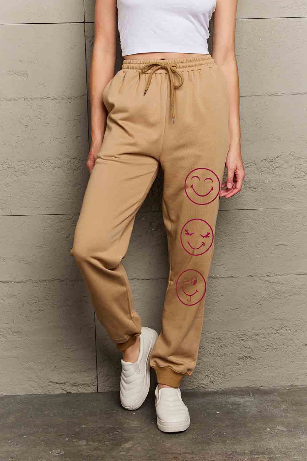 Woman wearing Graphic Sweatpants with emoji print and drawstring waist, standing against a concrete wall.