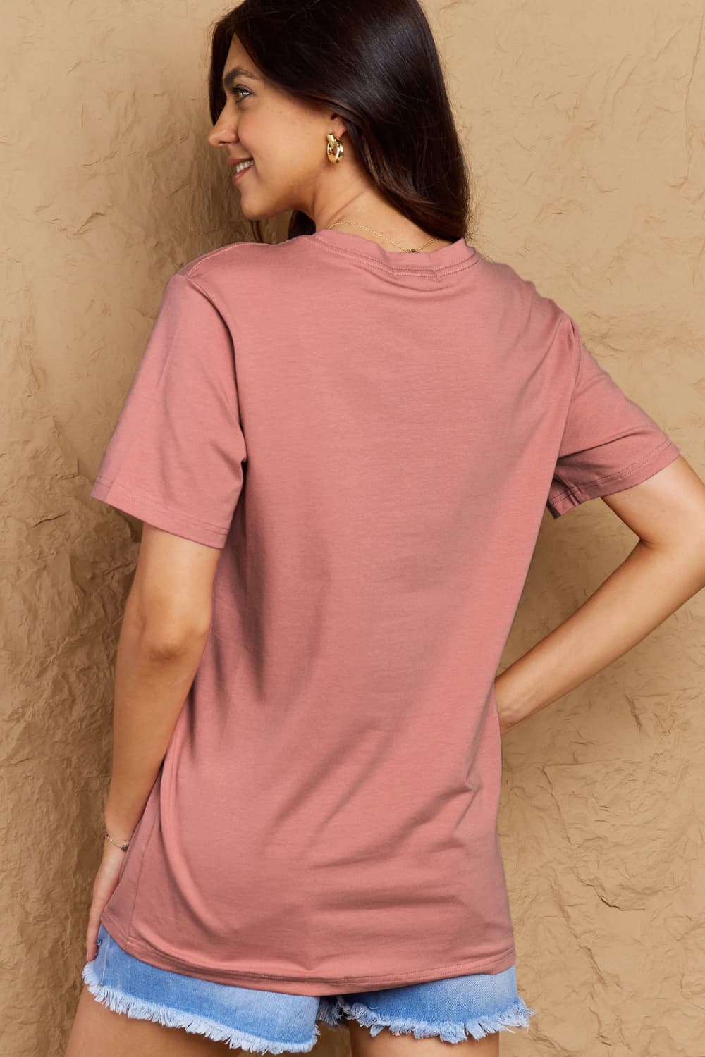 Woman wearing Graphic Cotton T-Shirt, FIND INNER PEACE print, back view, dusty rose color, paired with denim shorts.