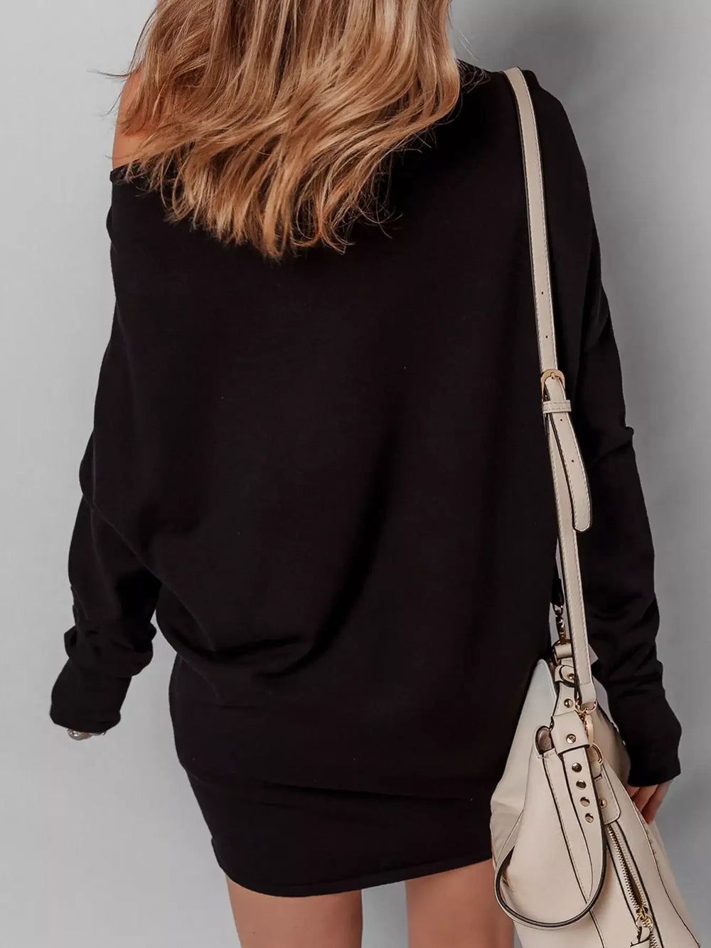 Back view of a woman wearing an asymmetric batwing sweater dress, featuring a mini length and off-shoulder design, paired with a white bag.